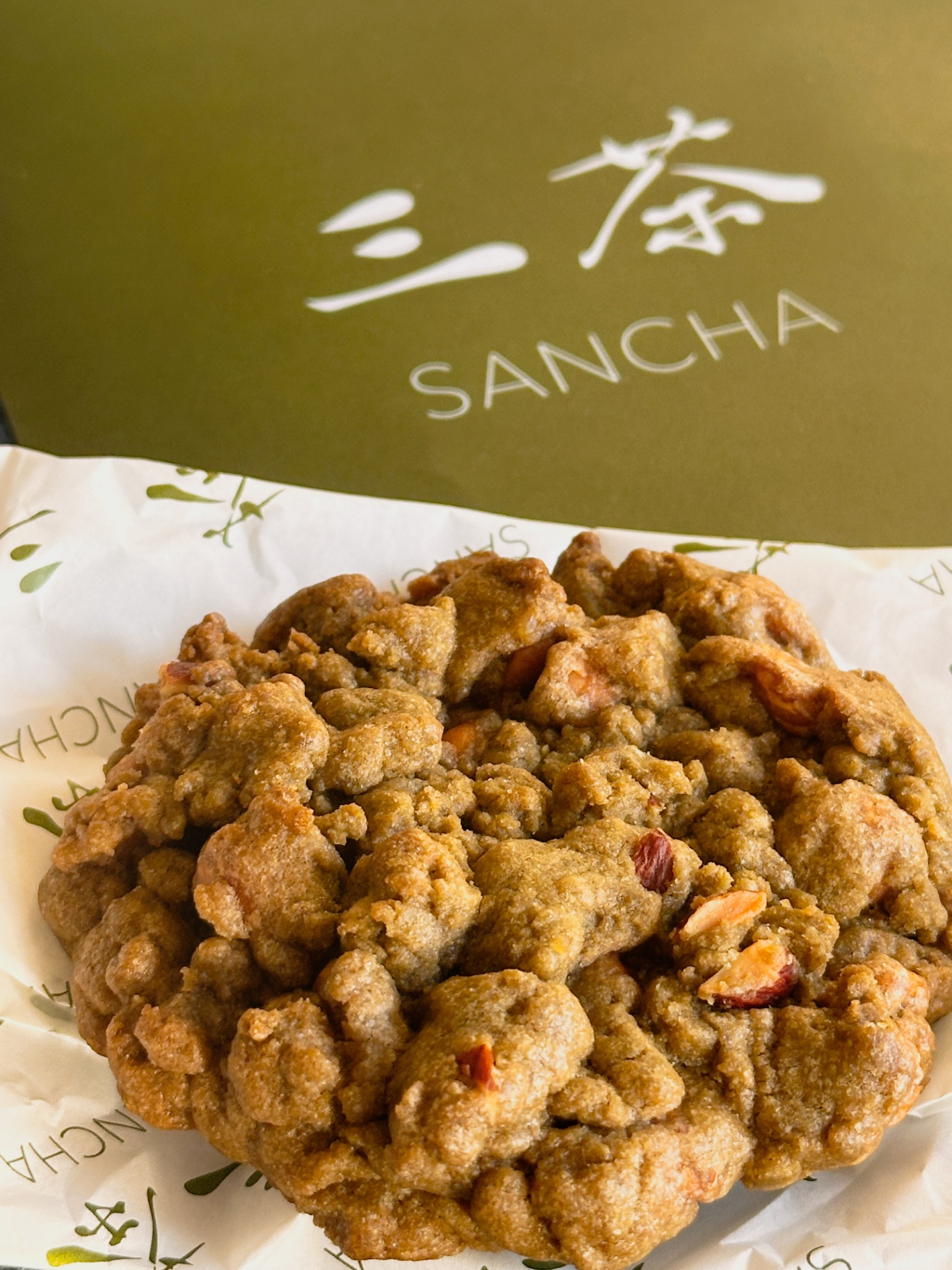 Close-up of a large, textured Sancha cookie with visible nuts, resting on branded paper in front of a matcha-green Sancha box