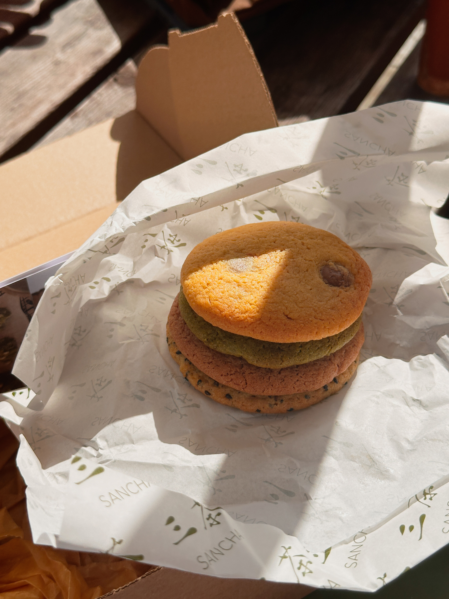 Stack of four Sancha cookies — kinako, matcha, strawberry, and black sesame — resting on branded paper