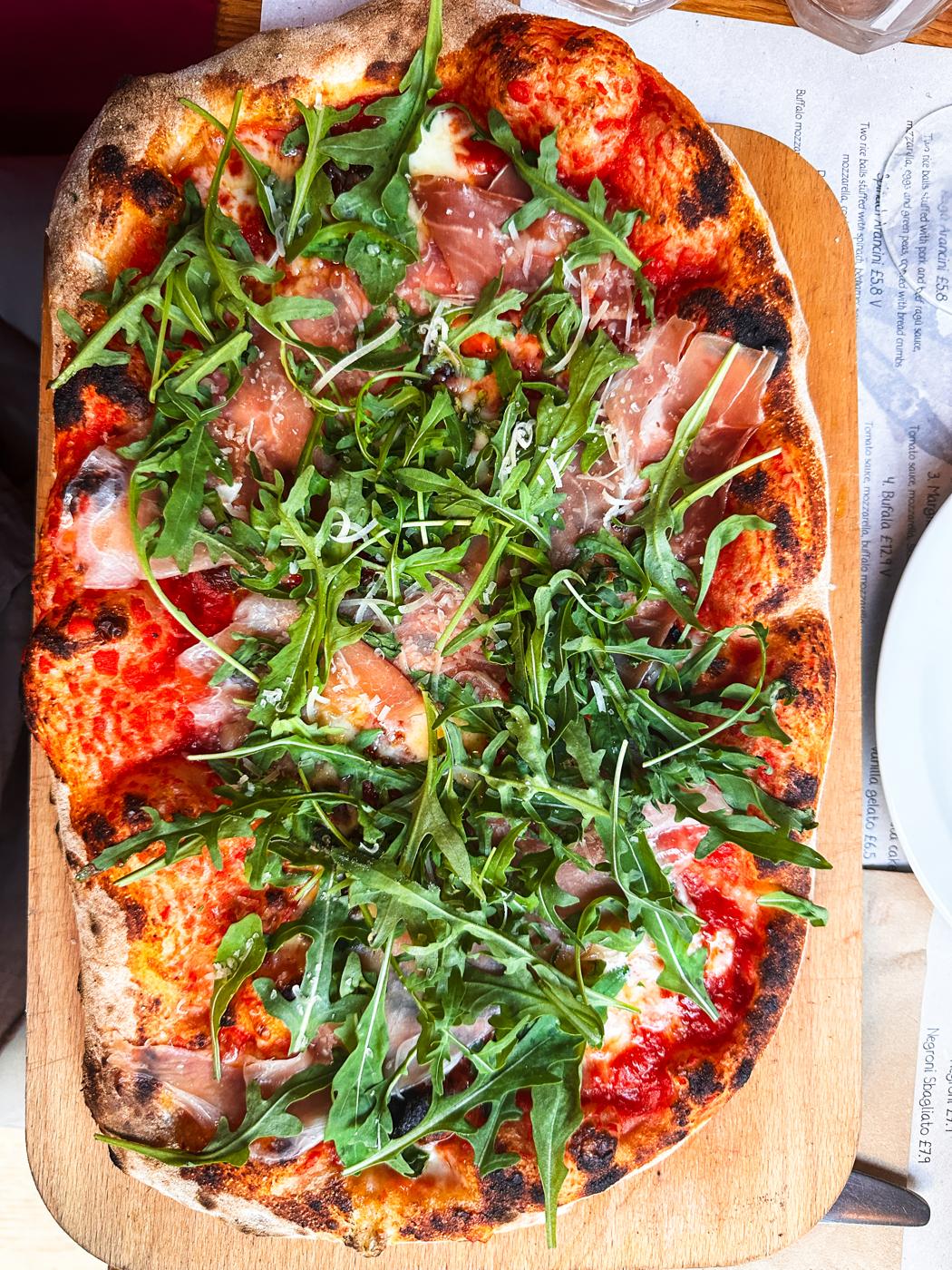 Sourdough pizza topped with rocket and cured ham on a wooden board at Aromi, Bene't Street, Cambridge